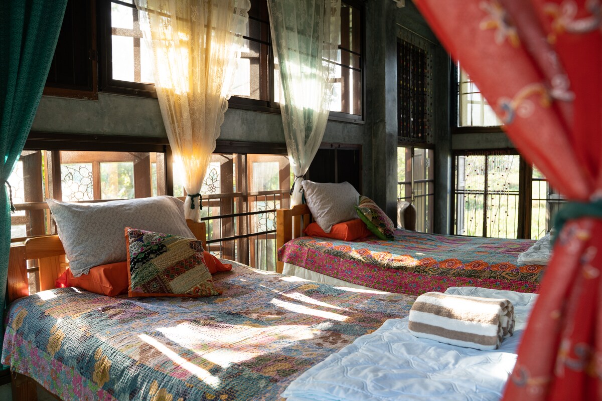 A bright room features two spacious beds adorned with colorful quilts, complemented by decorative cushions. Large windows allow natural light to fill the space, while light, airy curtains create a welcoming ambiance. Fresh towels are neatly arranged on a bed beside a traditional textile.