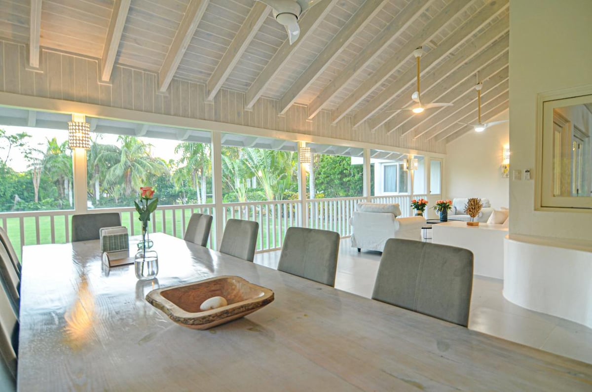 A spacious dining area features a large wooden table surrounded by light gray upholstered chairs. Natural light pours in through panoramic windows, showcasing views of the lush garden. Beautiful pendant lights hang from the ceiling, complementing the villa's open and airy design.