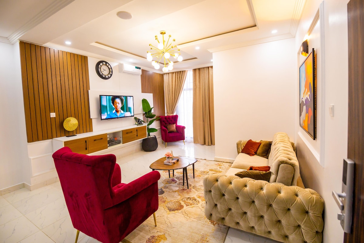The spacious living room features a plush, tufted sofa and two stylish armchairs in rich red hues. A sleek television is mounted on a modern wall unit adorned with decorative elements. Large windows are dressed with elegant curtains, allowing natural light to fill the space.