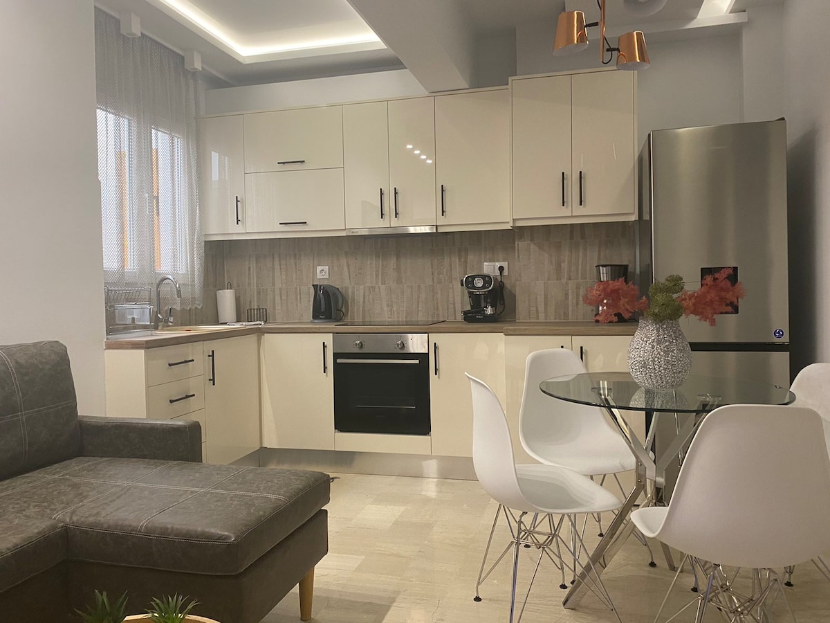 A modern kitchen area features sleek white cabinetry and stainless steel appliances, including a refrigerator and oven. A round glass dining table is surrounded by white chairs, while a corner sofa adds comfort to the open layout. Natural light enters through window treatments.
