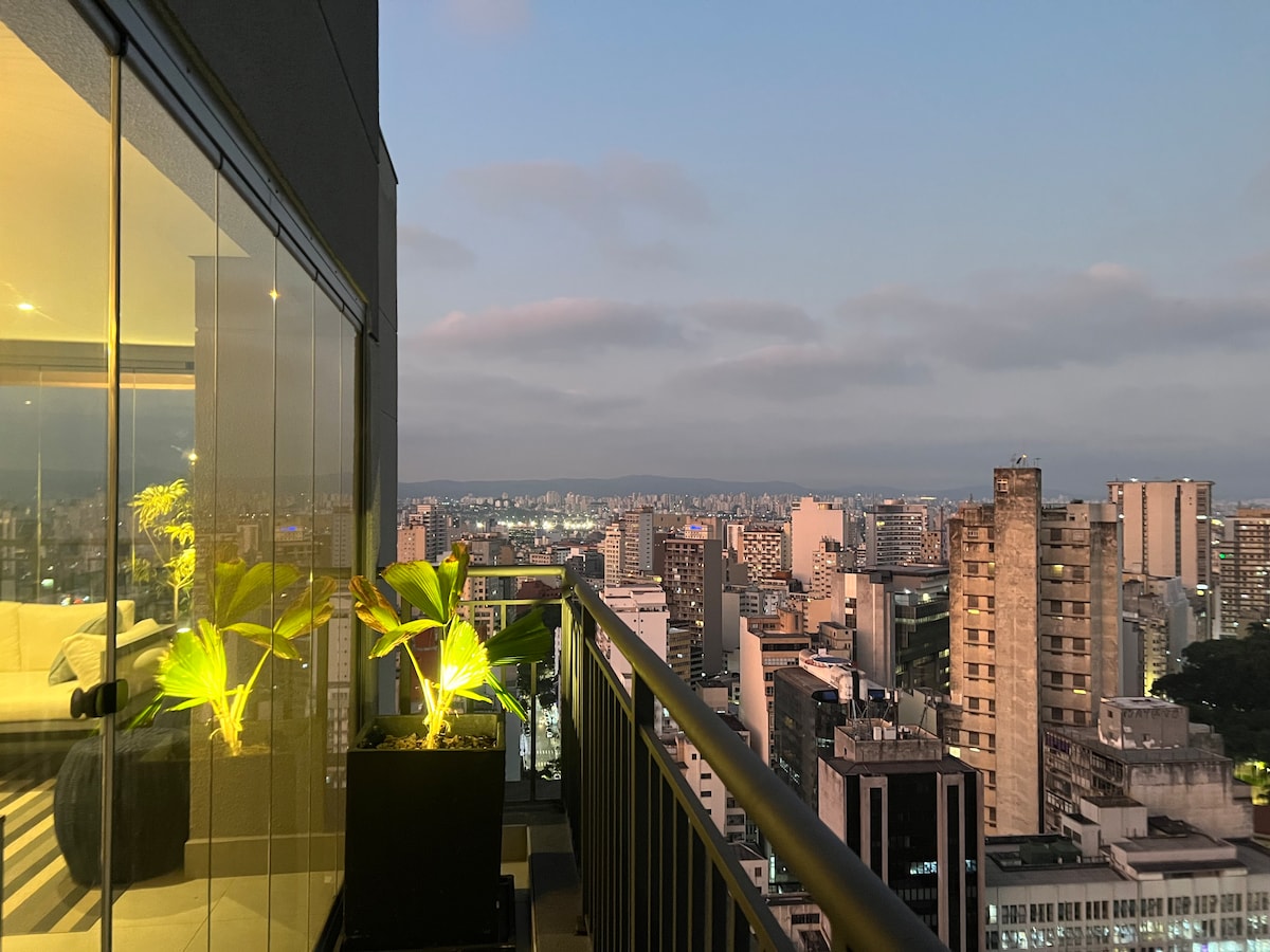 A balcony view features city lights and a skyline, with illuminated plants in stylish black planters. The glass railing allows for unobstructed sightlines of the surrounding buildings, creating a serene space to enjoy the urban landscape.