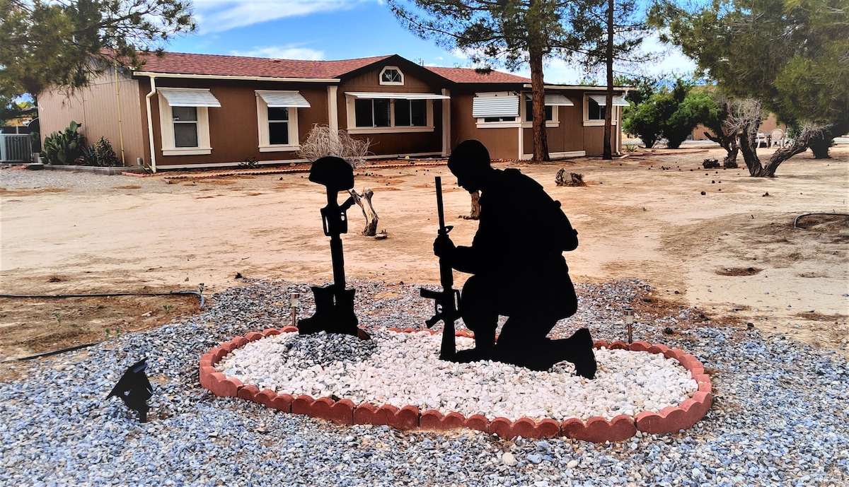 A memorial sculpture is displayed in a gravel bed, featuring two silhouettes of soldiers with rifles, kneeling in a respectful position. A single-story house with large windows is visible in the background, surrounded by desert landscaping and a clear blue sky.