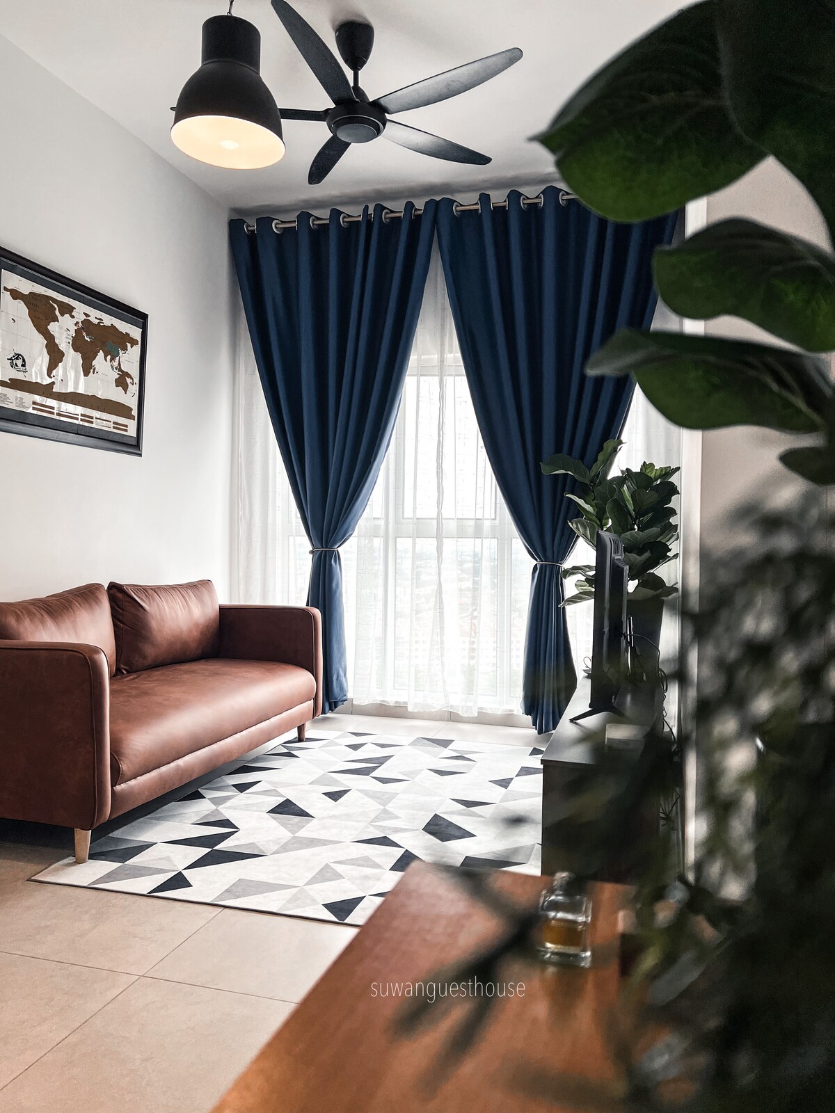 A modest living area is presented featuring a brown sofa positioned against a wall. Large blue curtains frame a window that allows natural light to fill the space. A geometric patterned rug lies on the floor, complemented by indoor plants and wall art.