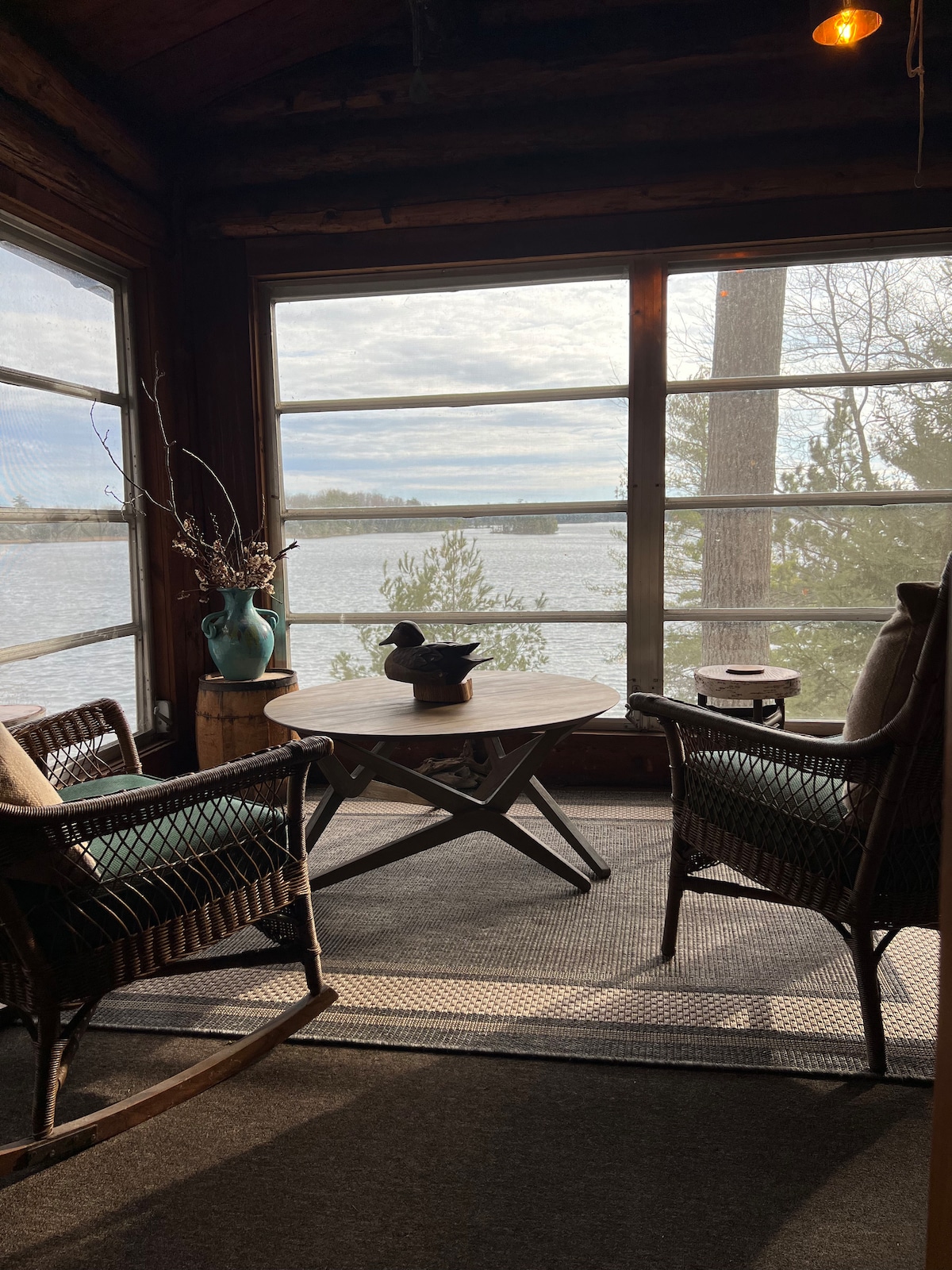 A cozy seating area is presented with two wicker chairs placed around a small wooden table. Large windows offer a view of the lake and surrounding nature, allowing natural light to fill the space. Decorative elements, including a vase and a small decorative duck, enhance the ambiance.