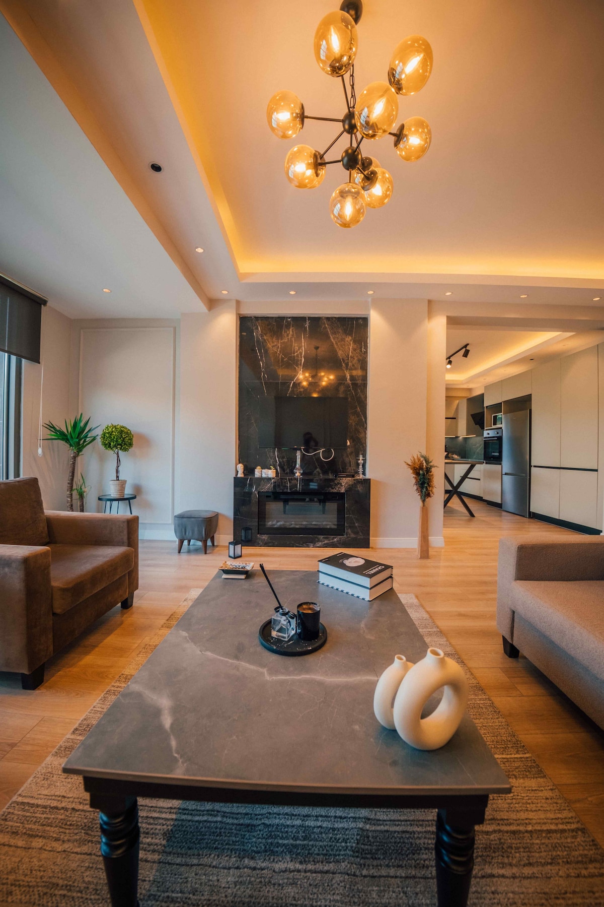 A spacious living area features natural light streaming in through large windows. Elegant furniture, including two armchairs and a coffee table with a marble top, is present. An ornate chandelier hangs from the ceiling, while a modern fireplace adds a touch of sophistication.