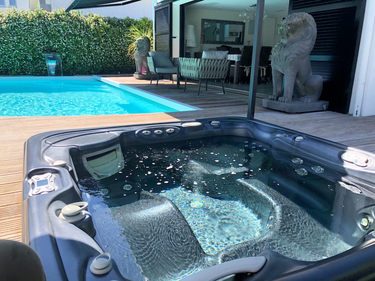 A private spa with multiple jets is situated near a glistening pool. The outdoor area features stylish seating arrangements under a shaded canopy, with greenery providing privacy. Decorative statues add an elegant touch to the inviting space.
