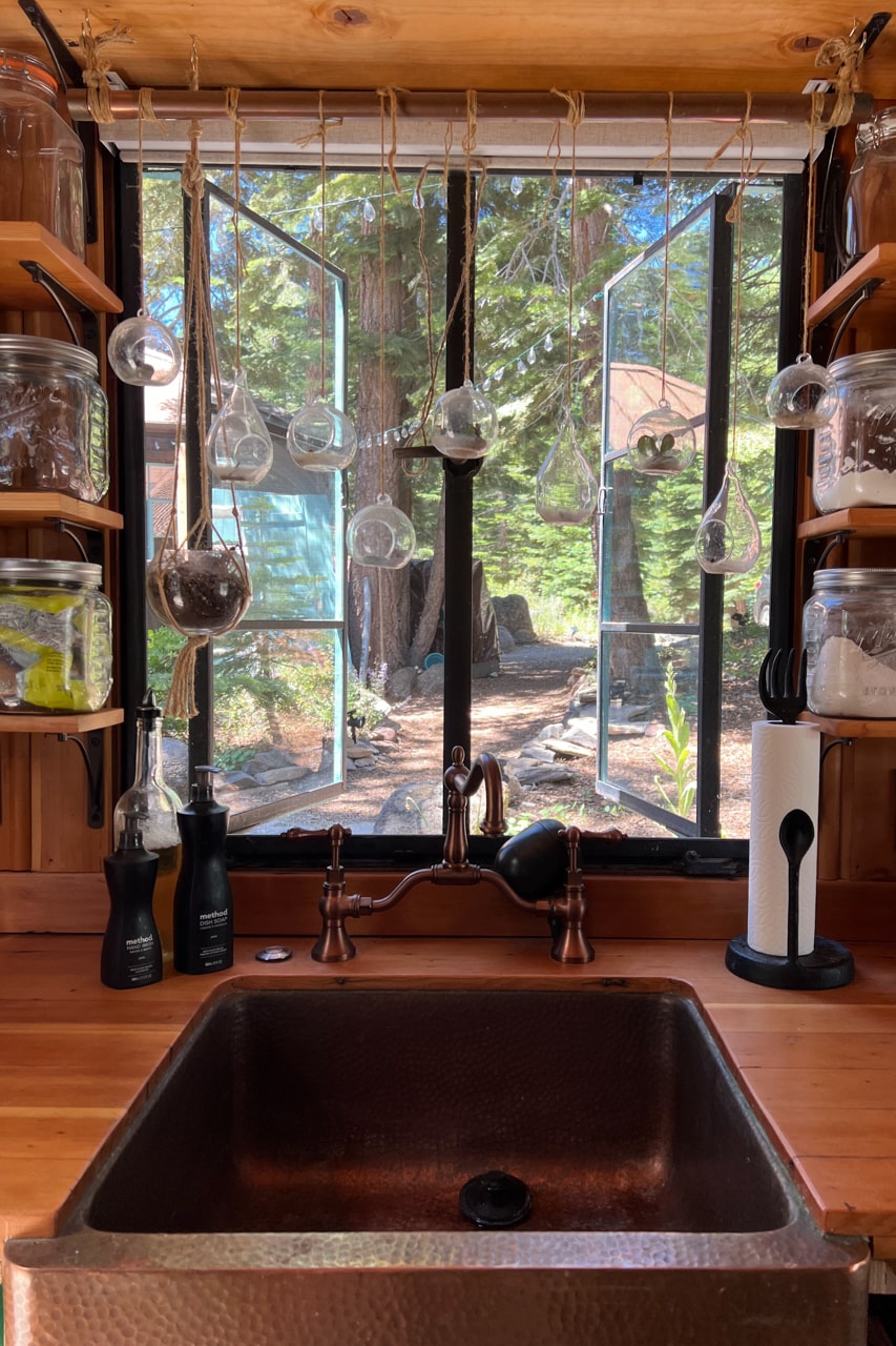 A well-lit kitchen sink area is featured, with a copper basin beneath a window framed by wooden shelves displaying glass jars. Sunlight filters through, illuminating hanging glass ornaments and showcasing the lush greenery outside.