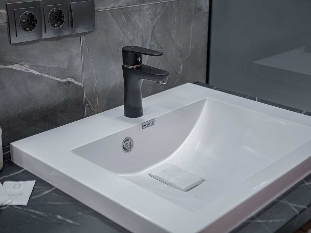 A modern bathroom sink is positioned on a sleek dark marble countertop. The square-shaped white basin features a single elegant faucet with a matte black finish, surrounded by minimalist wall outlets.