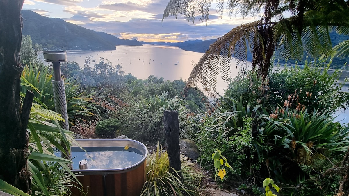 A wood-fired outdoor bathtub is nestled among lush greenery, offering a serene view of the water below. The tranquil landscape features distant hills and is framed by ferns, capturing the essence of nature's beauty during sunset.