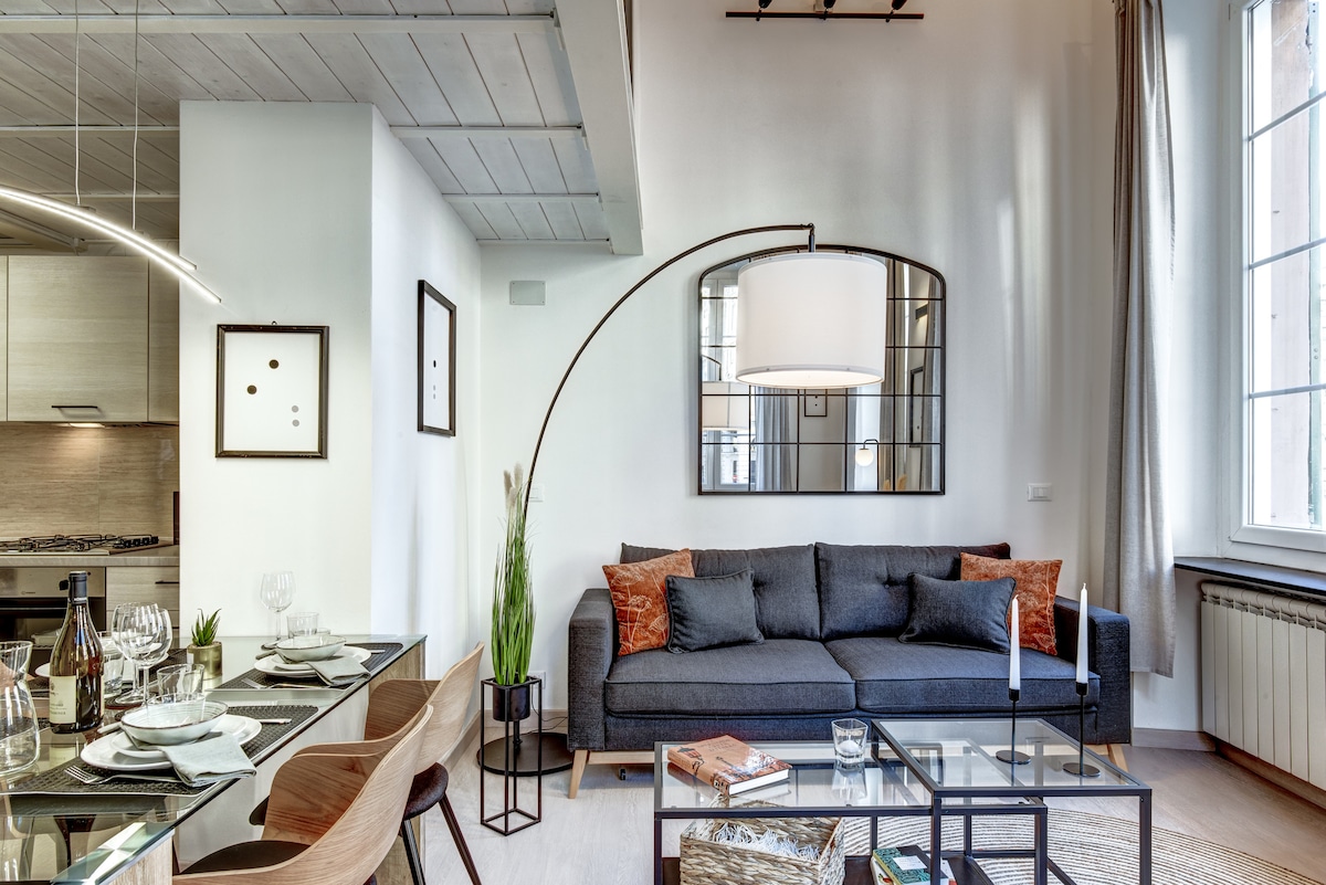 A stylish open living area features a modern gray sofa paired with decorative pillows, illuminated by a sleek floor lamp. A glass dining table is set for four, positioned next to large windows that fill the space with natural light.