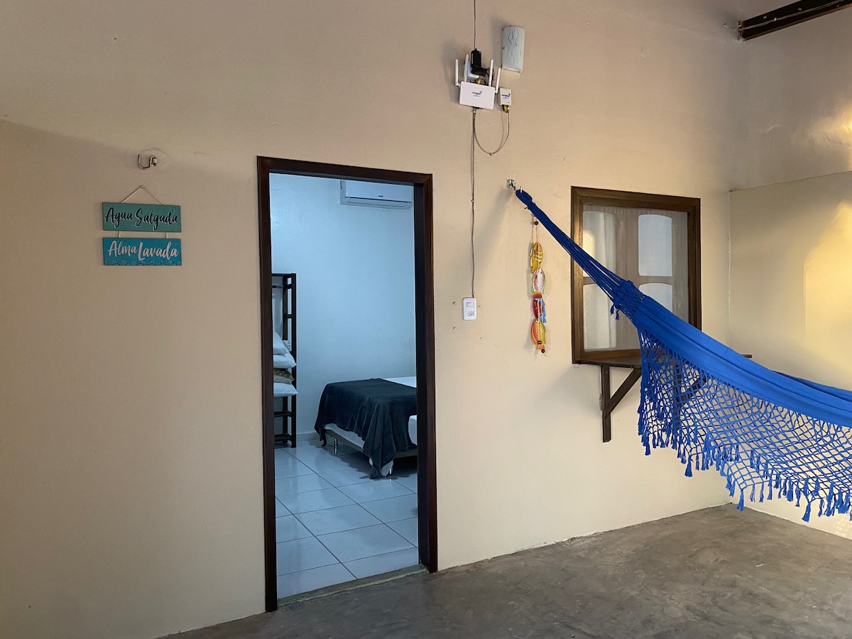 An indoor space features a colorful hammock hanging beside a window. A doorway leads to a separate sleeping area, which contains a spacious bed with light-colored linens. A small sign on the wall indicates the room's name, adding a personal touch.