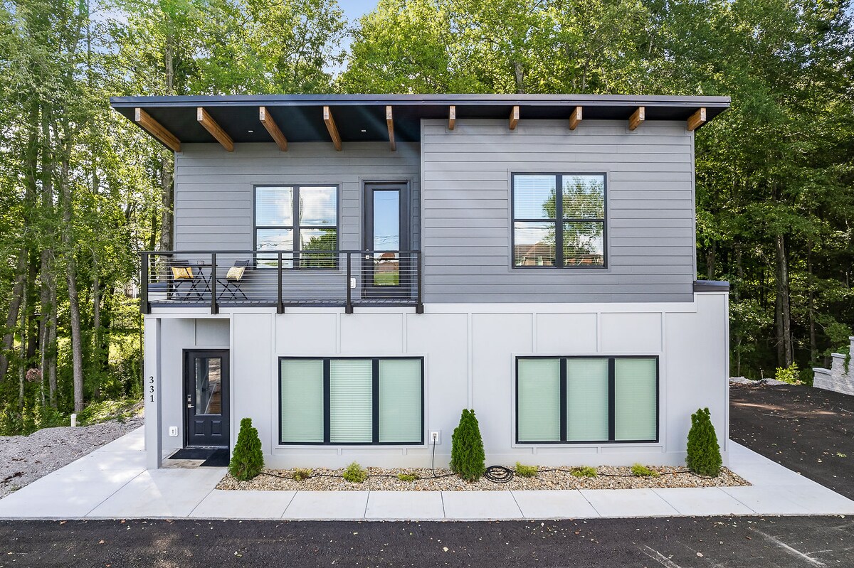 The exterior of the modern two-story building is showcased, featuring a combination of gray siding and white walls. Large windows provide ample natural light, while the covered balcony highlights outdoor seating. Lush greenery surrounds the property, enhancing the inviting atmosphere.