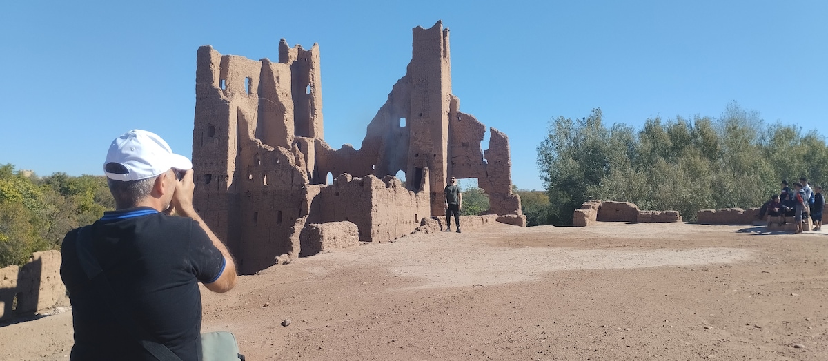 An ancient earthen structure is portrayed, showcasing remnants of walls and arches against a clear blue sky. Visitors are seen exploring the site, with lush greenery in the background. The ground appears dusty and natural, enhancing the historic ambiance of the location.