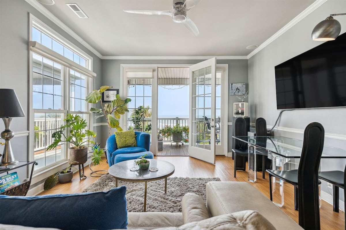 A bright living area is showcased, featuring large windows that invite natural light. French doors open to a balcony with ocean views. A blue chair and green accents complement stylish furnishings, alongside a glass table and a modern entertainment unit mounted on the wall.