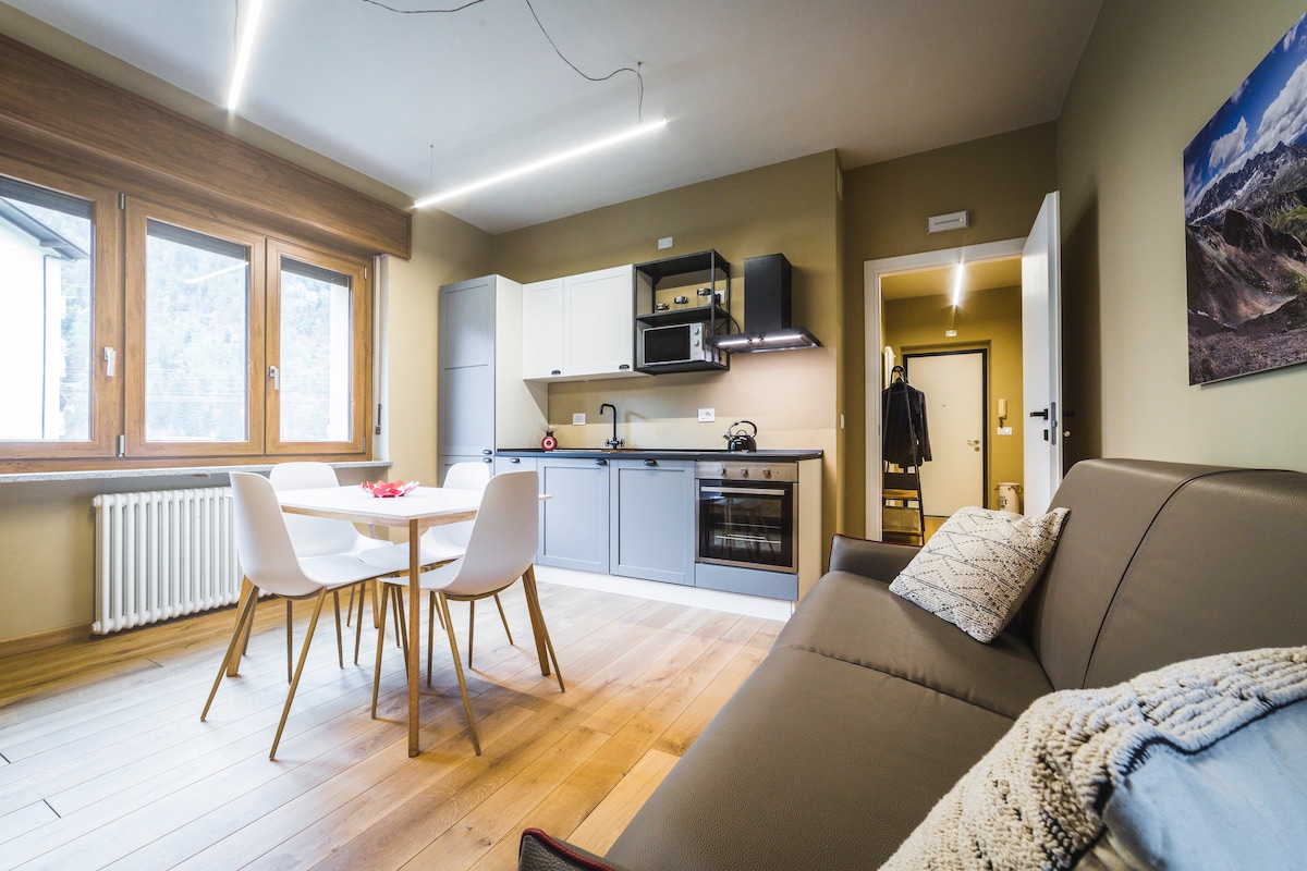 A modern kitchen and dining area features a light wood floor. An inviting dining table is surrounded by white chairs, while gray cabinetry complements stainless steel appliances. Natural light filters through large windows, illuminating the open space. A cozy sofa is positioned nearby.