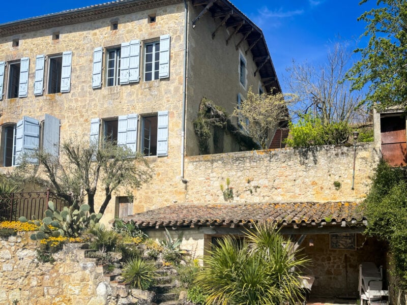 The stone exterior of a large village house is visible, displaying traditional blue shutters and surrounded by landscaped gardens. Lush greenery, including palm and cactus plants, complements the rustic stone wall and terrace area, with glimpses of the clear blue sky overhead.