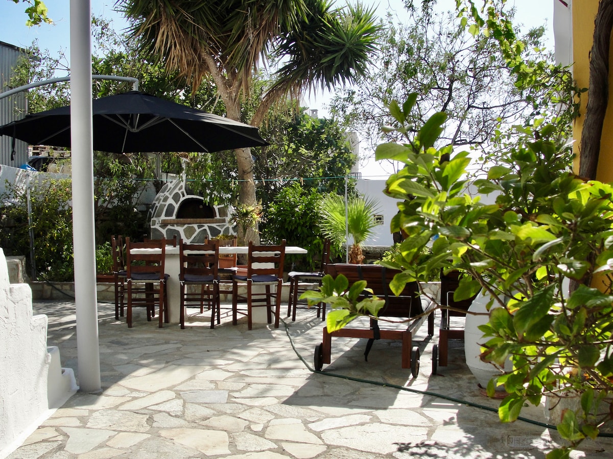 A private courtyard is surrounded by lush greenery, featuring a stone-paved area with wooden tables and chairs. An umbrella provides shade over the seating area, while a barbecue is visible in the background, enhancing the outdoor space for relaxation and dining.