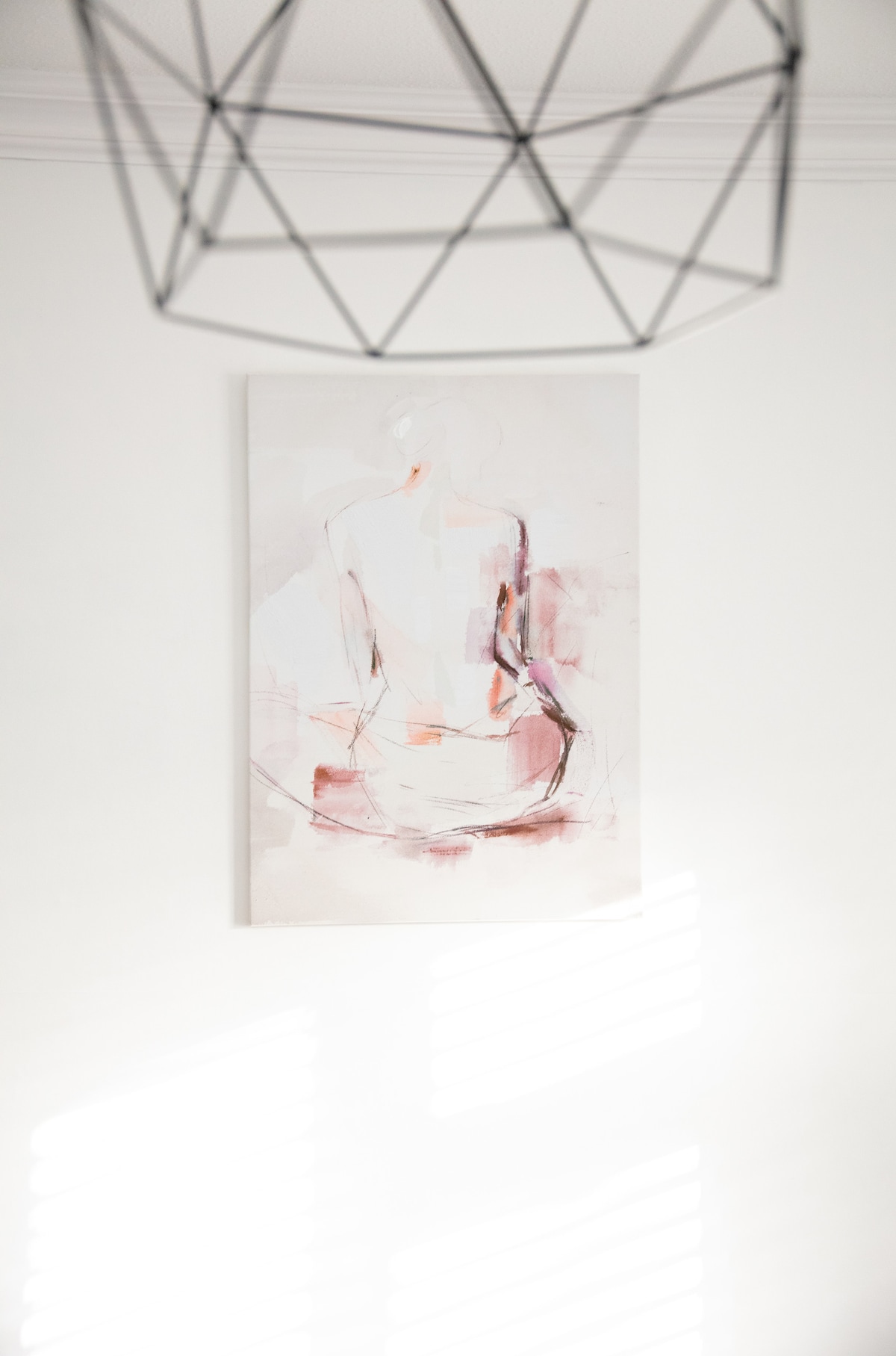 An abstract painting is displayed on a white wall, featuring a softly rendered figure with hues of pink and subtle strokes. Natural light creates a gentle interplay of shadows across the artwork, enhancing its delicate appearance.