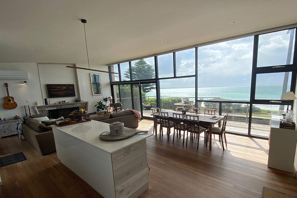 A spacious living area is shown, featuring modern furnishings and a well-equipped kitchen. A large dining table accommodates multiple guests, while expansive wall-to-ceiling windows offer uninterrupted views of the coastline and the ocean beyond.