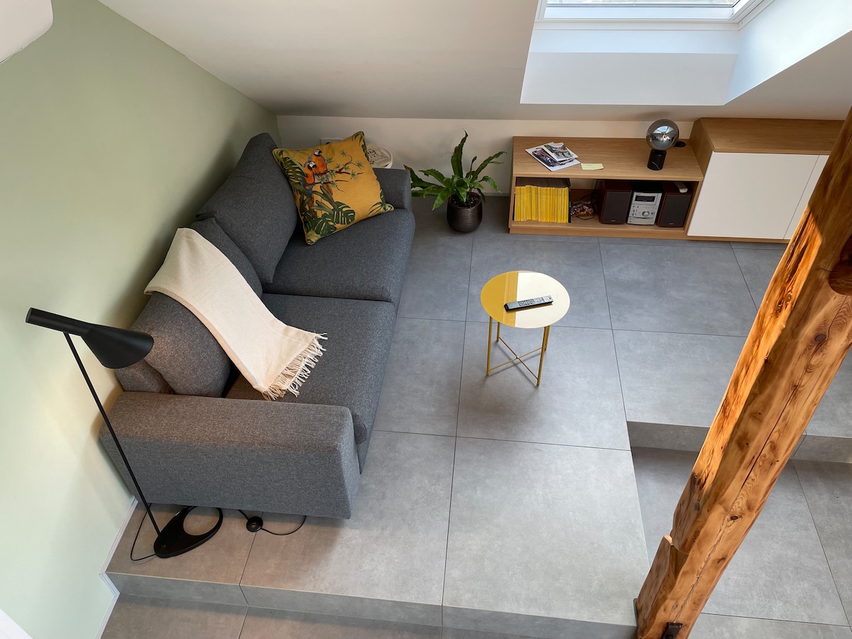 A comfortable gray sofa is situated in a well-lit corner, accompanied by a decorative pillow and a soft throw. A small round gold table sits nearby on tiled flooring. A potted plant adds a touch of greenery, while natural light floods in through the skylight.