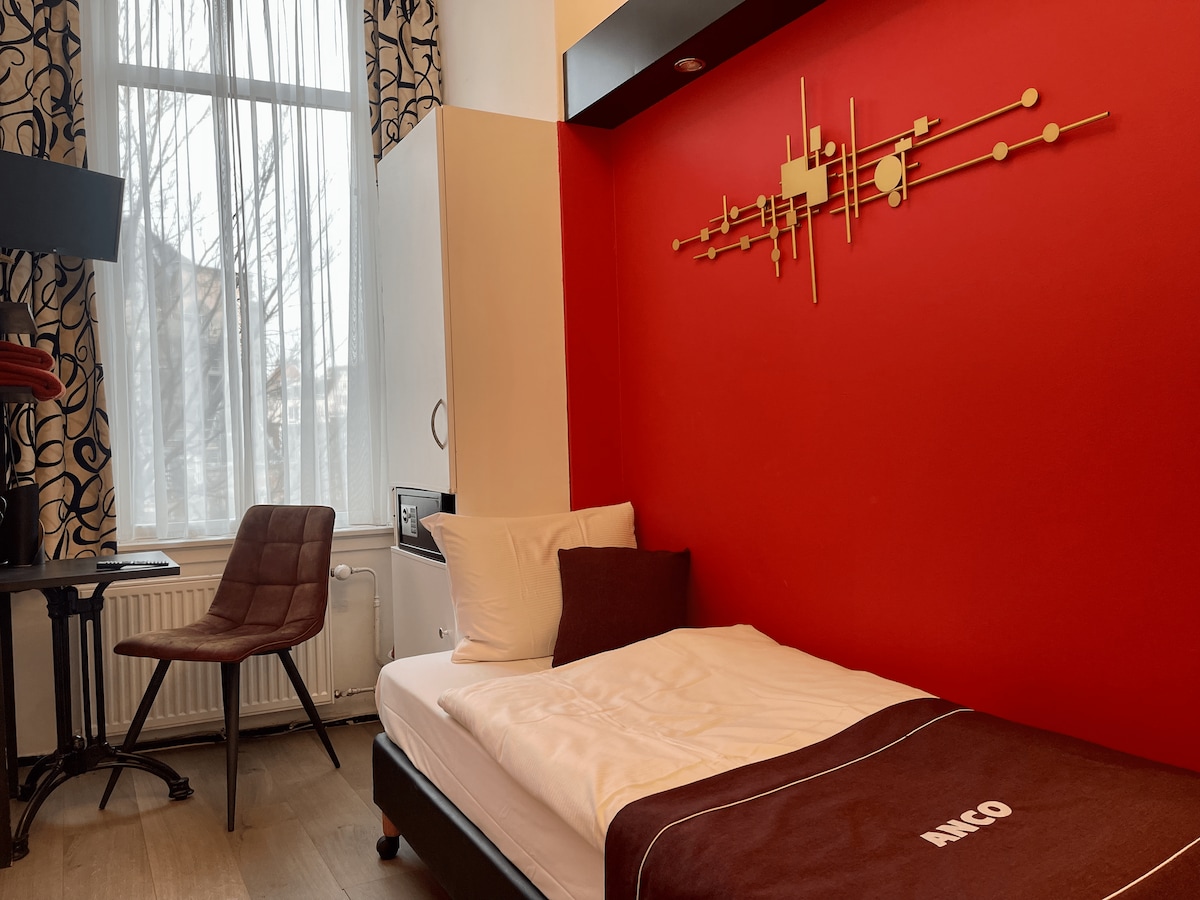 A compact sleeping area features a single bed dressed in crisp white linens and a dark brown throw. A striking red wall serves as a backdrop, accented by modern wall art. A small desk and chair are positioned near a window, allowing natural light to enter.