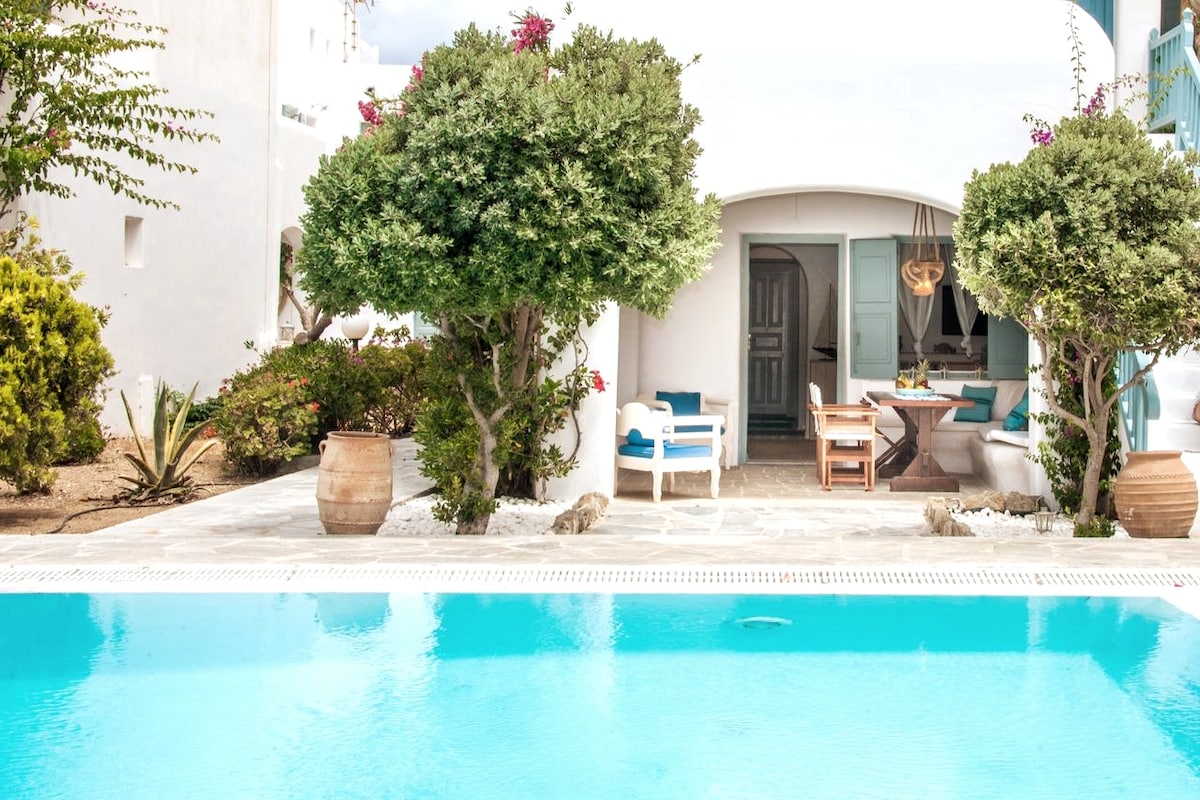 A serene outdoor area showcases a large swimming pool surrounded by neatly arranged plants. Two trees provide shade, complementing comfortable seating options nearby. A wooden table and chairs are positioned in front of a white-walled building with blue accents, inviting relaxation and gatherings.