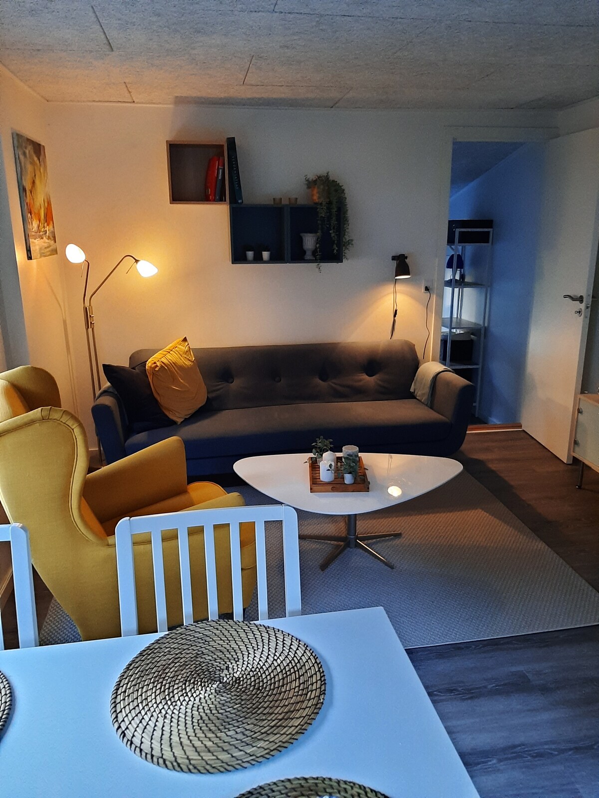 A bright living area is visible, featuring a gray sofa with colorful accent pillows. A white coffee table is placed in front, surrounded by yellow armchairs. A cozy dining table is set with woven placemats, and an open bookshelf adds a decorative touch to the space.