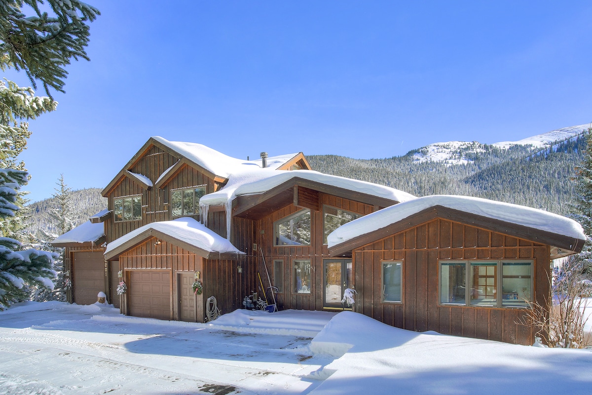 The exterior of a contemporary log cabin is presented, featuring a mix of brown wood planks and expansive windows. The structure is set against a backdrop of snow-covered mountains, with a serene winter landscape and a clear blue sky overhead.