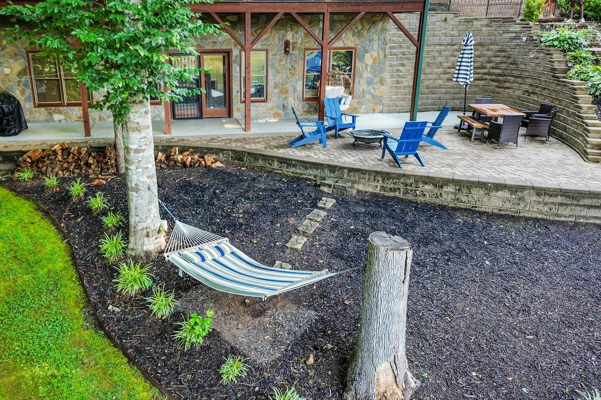 A serene outdoor space features a striped hammock suspended between two trees, inviting relaxation. Nearby, blue Adirondack chairs are arranged around a fire pit on a stone patio, surrounded by thoughtfully landscaped grounds with various shrubs and flower beds.