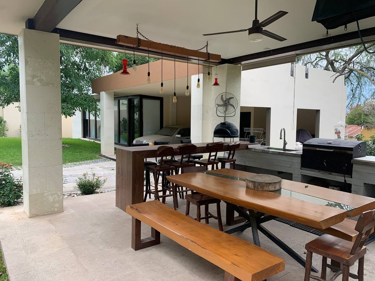 An outdoor dining area features a long wooden table surrounded by benches, complemented by a bar with high stools. A grill is placed nearby, and ceiling fans are installed for comfort. Verdant landscaping can be seen in the background, enhancing the outdoor experience.