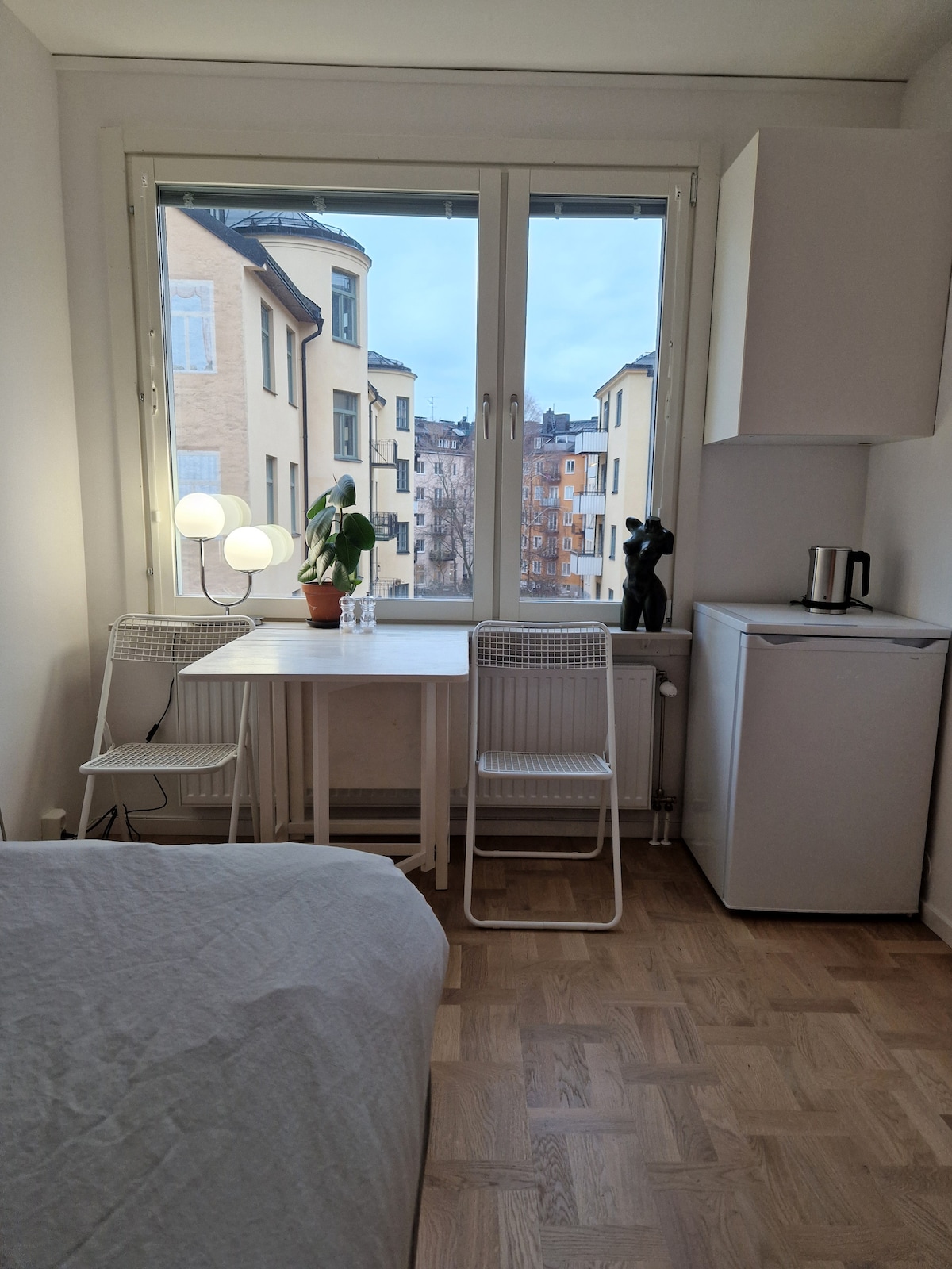 A small dining area features a table accompanied by two chairs, positioned near a large window. A potted plant and a sculpture add visual interest. The compact refrigerator and kettle are situated to the right, emphasizing the efficient layout of the space.