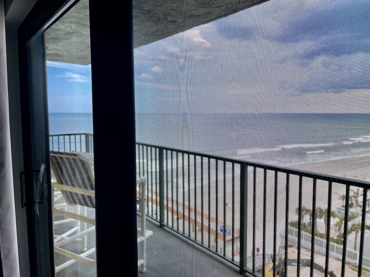 A balcony view showcases the ocean, with gentle waves lapping the shore. A sun lounger is positioned on the balcony, allowing for relaxation. Beyond the railing, the beach and a few palm trees are visible, creating a serene coastal scene.
