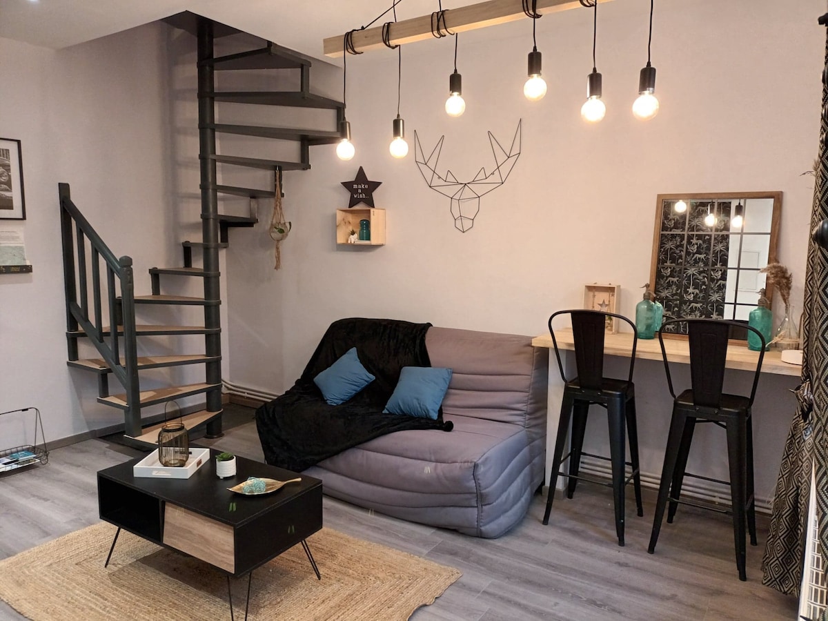 A modern living area features a grey sofa with blue cushions and a decorative deer head on the wall. A spiral staircase ascends to the upper level. A wooden coffee table sits on a natural fiber rug, complemented by bar stools and a small desk with decorative items.