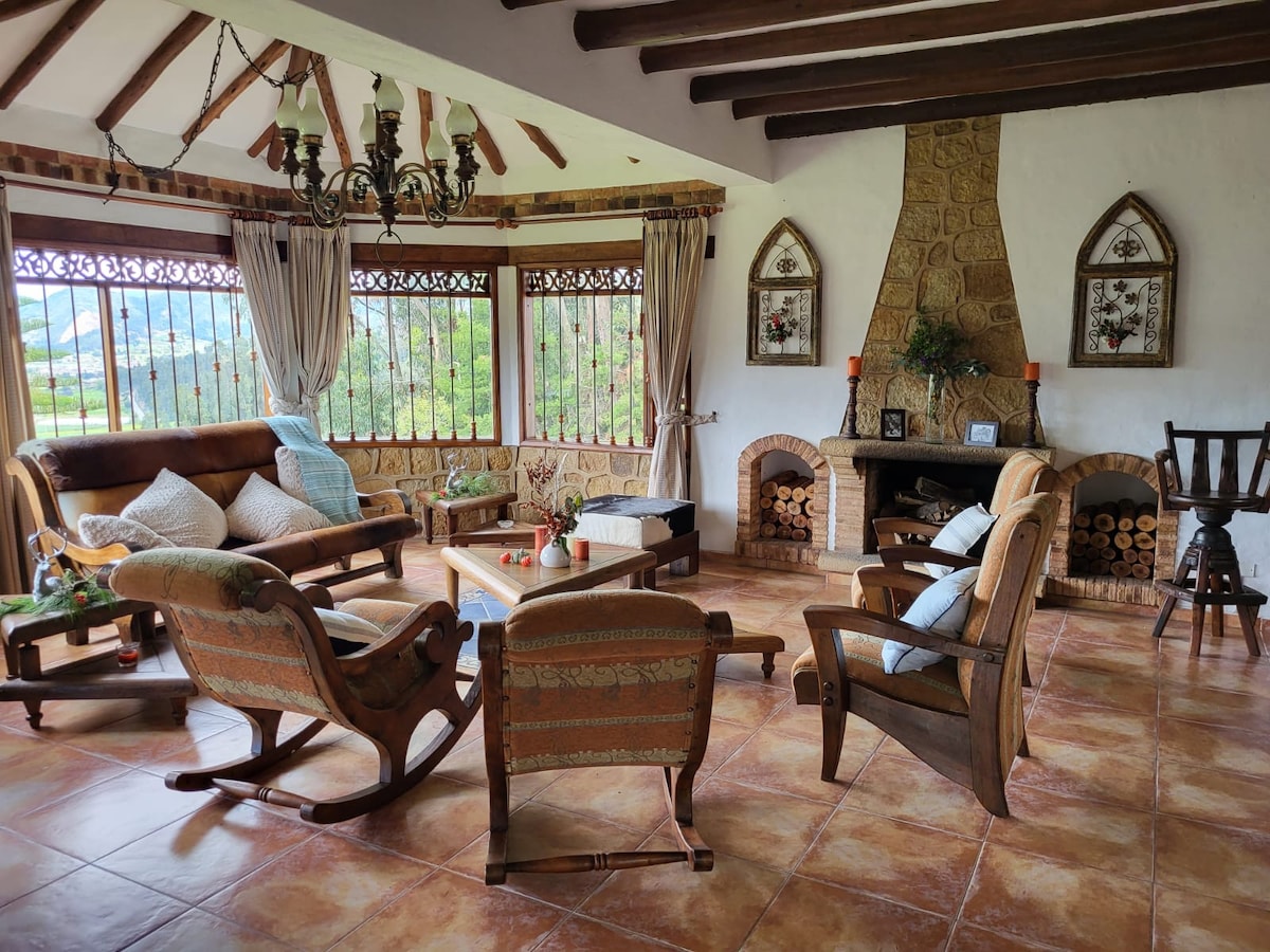 A spacious living area features wooden beams and large windows that frame panoramic countryside views. Comfortable seating options, including rocking chairs and a sofa, surround a central coffee table. A stone fireplace is positioned against the wall, enhancing the cozy ambiance.