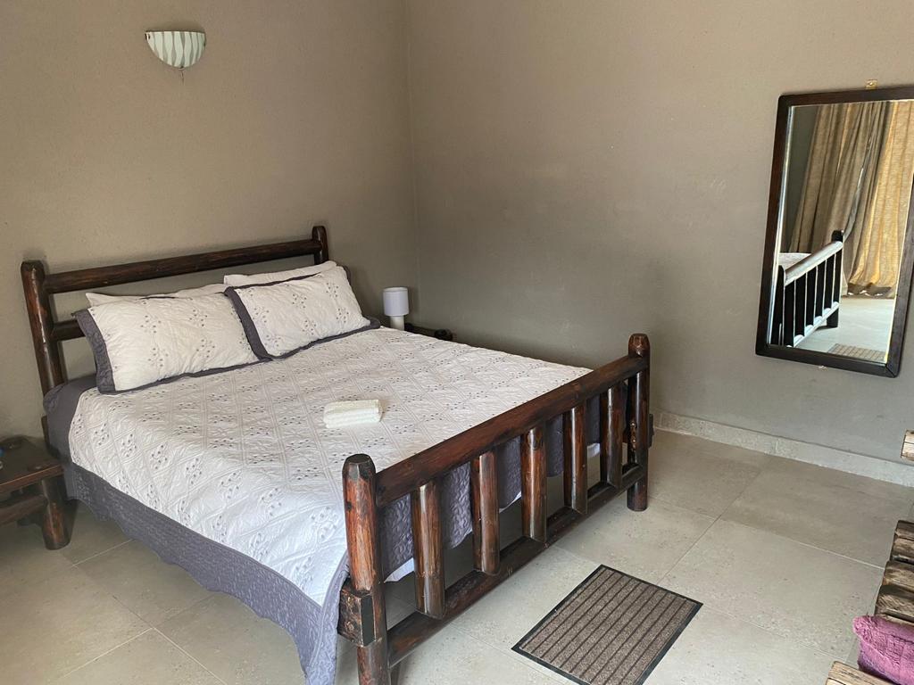 A wooden bed with a white comforter is centered in the room, accompanied by a bedside lamp. A large mirror reflects the space, and neutral-colored walls create a calm atmosphere. Light enters from a window covered by soft drapes.