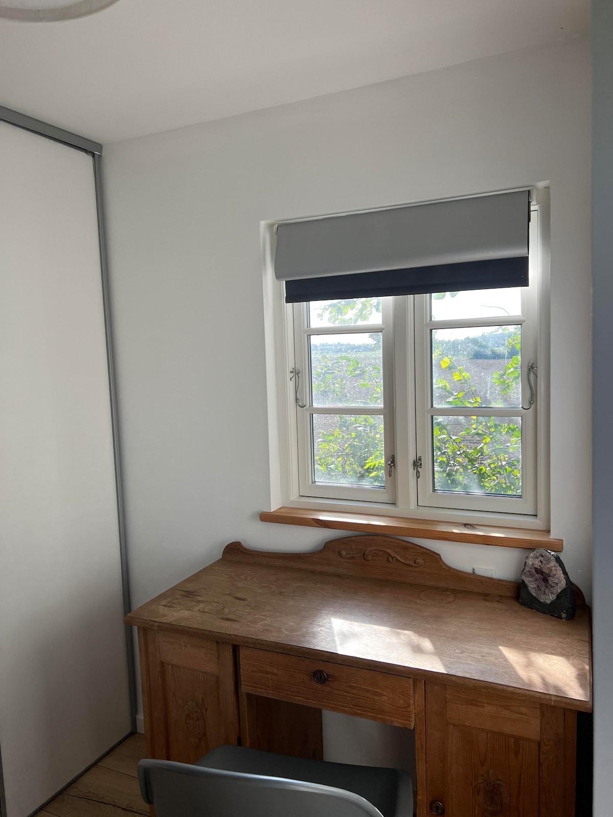 A wooden desk is positioned beneath a window that features a two-panel design with gray shades. Natural light filters through, illuminating the desk and highlighting the view of greenery outside. The walls are painted in light tones, enhancing the serene atmosphere.