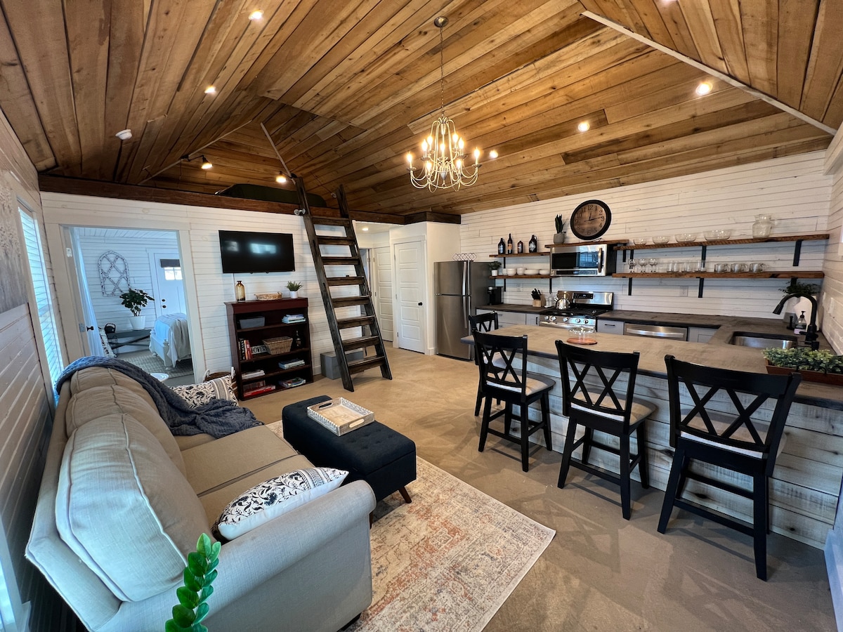 An open living area features a cozy couch and a stylish coffee table, with a modern kitchen visible in the background. A wooden staircase leads to an open loft, and a dining area with black chairs surrounds a sizable table. Contemporary lighting complements the rustic wood ceiling.