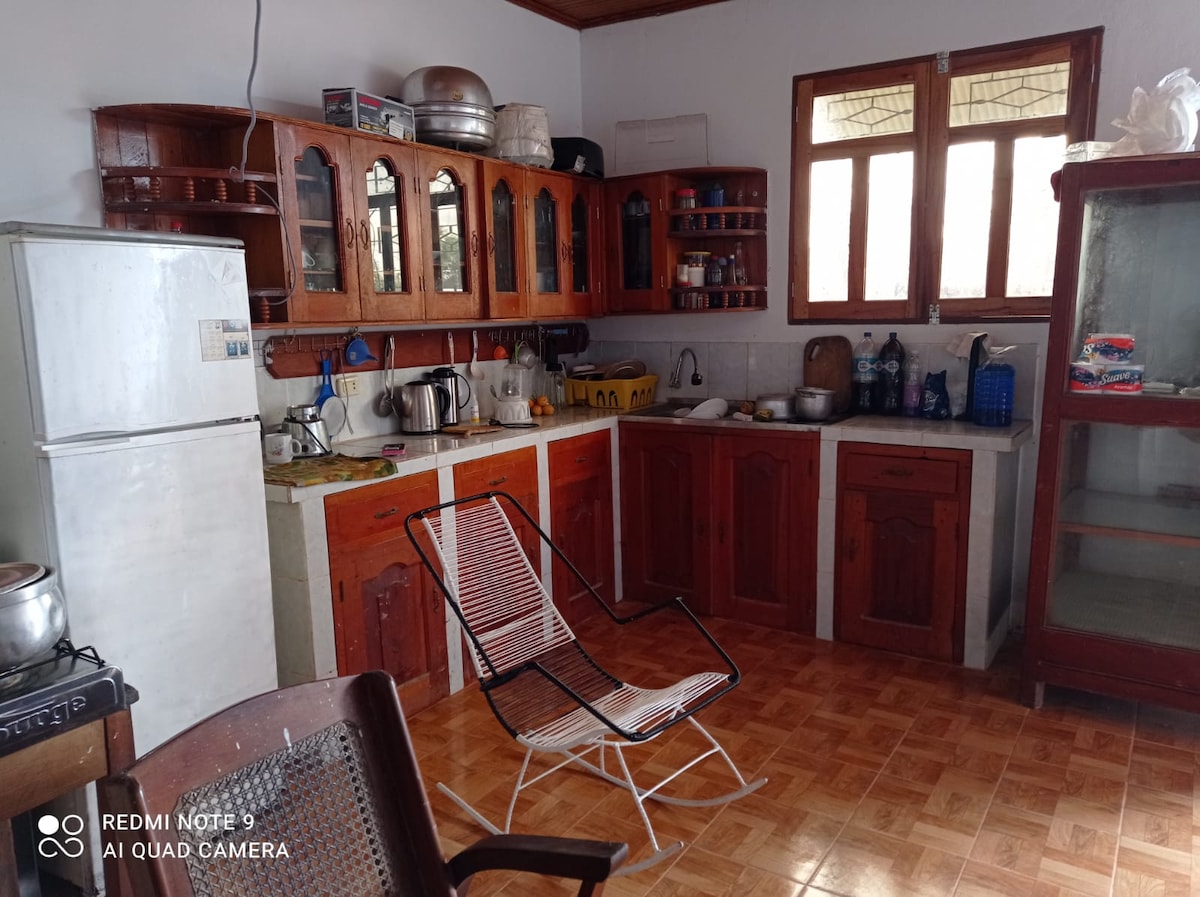 A functional kitchen features wooden cabinetry that provides ample storage. A refrigerator, stove, and various kitchen appliances are visible. A small table is positioned near a window, allowing natural light to enhance the space. A folding chair is placed nearby for convenience.