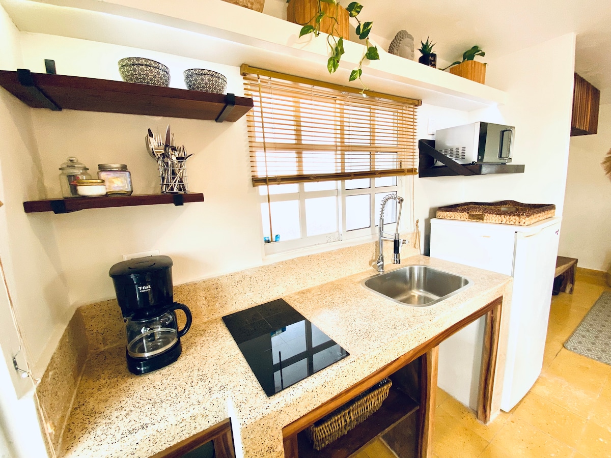 A functional kitchen space is presented, featuring a counter with a sink and stovetop. Open shelving displays kitchen utensils and decorative items. A coffee maker and microwave are included, alongside a compact refrigerator. Natural light filters through a window adorned with wooden blinds.