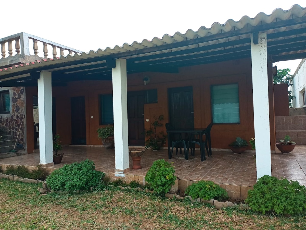 A covered outdoor patio features a tile floor, complemented by potted plants and greenery. A small table and chairs are arranged for seating. The entrance to the home is visible, showcasing wooden doors and large windows that allow natural light.