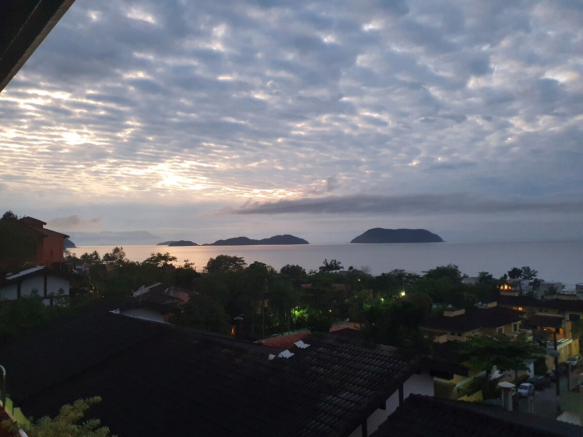 A panoramic view of the ocean is captured under a cloud-covered sky, with faint outlines of distant islands visible. The scene is accentuated by soft, muted colors as the sun begins to rise, creating a serene coastal atmosphere surrounded by lush greenery.