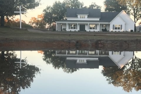 A two-story cabin is reflected in a calm pond, surrounded by trees. The exterior features a light-colored facade with a porch, offering a serene view. The tranquility of the water enhances the natural setting.