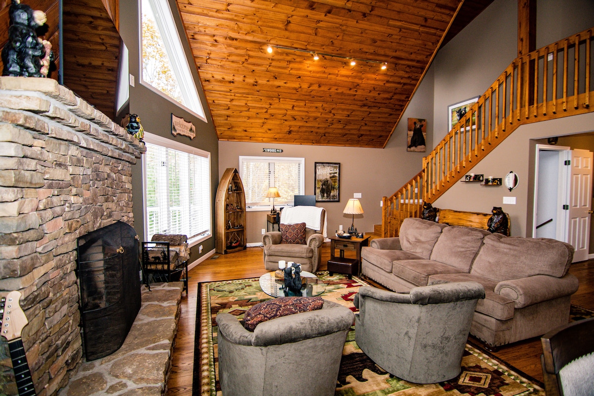 A spacious living area features a high vaulted ceiling with exposed wooden beams. Comfortable seating arrangements include a large sofa and two accent chairs positioned around a decorative area rug, with a stone fireplace serving as a focal point.