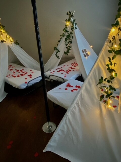 Two cozy sleeping areas are created with white fabric canopies, adorned with greenery and fairy lights. Petals are scattered across the beds, enhancing the inviting setup. A central pole supports the canopies, and warm lighting casts a soft glow in the space.