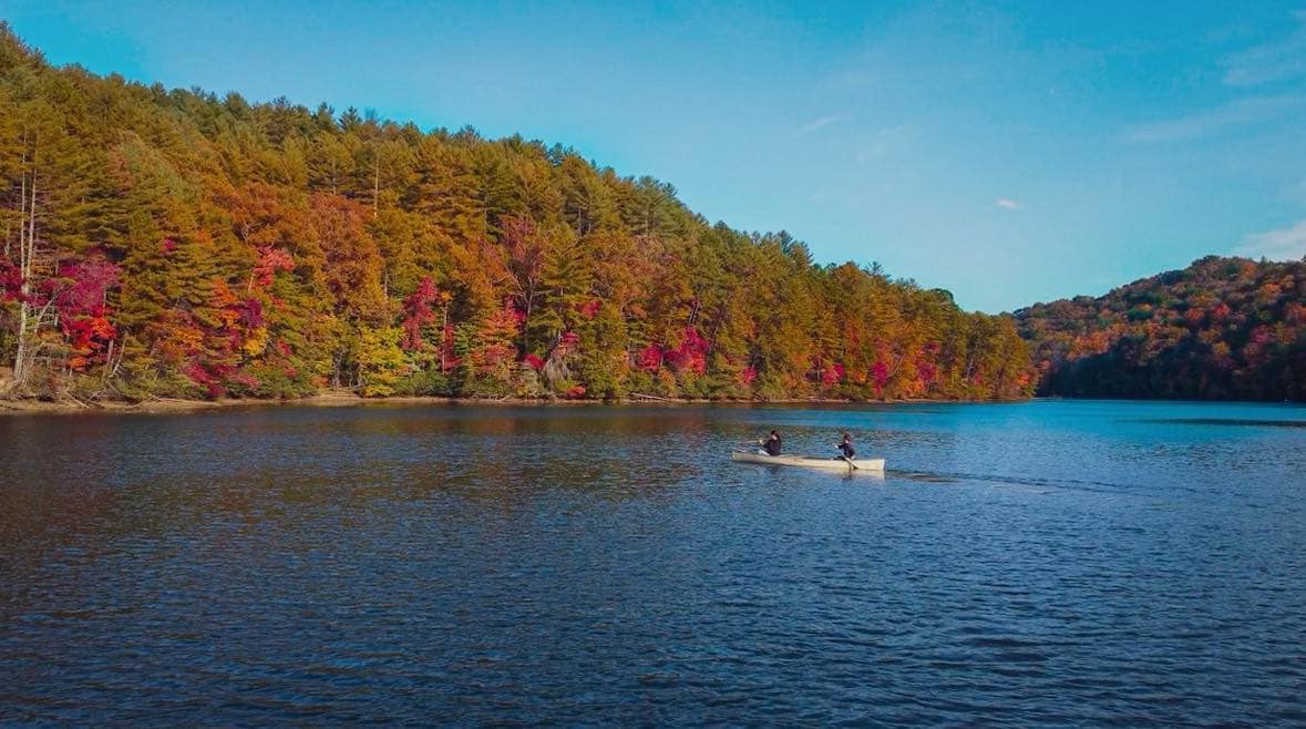 A serene lake scene is depicted, featuring two individuals in a canoe gliding across the calm water. Vibrant fall foliage surrounds the lake, with trees displaying rich hues of red, orange, and yellow. A clear blue sky frames the picturesque landscape.