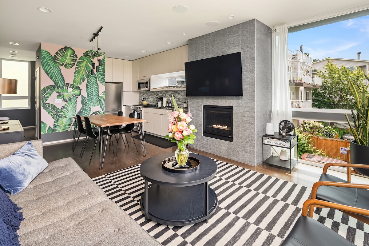 A modern living area features large windows allowing natural light to fill the space. A cozy seating arrangement surrounds a round coffee table, complemented by a stylish dining table nearby. Green foliage wallpaper adds a vibrant touch, alongside a wall-mounted television and a contemporary fireplace.