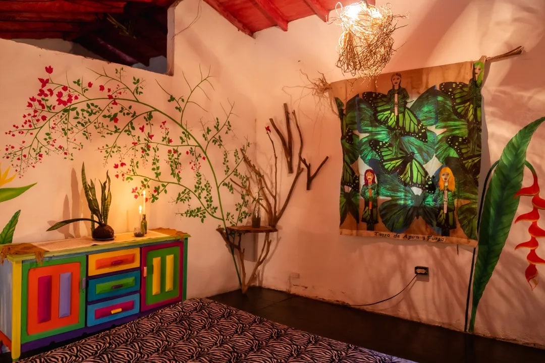 A vibrant room features a handmade, colorful cabinet and a decorative wall tapestry depicting figures in a lush green setting. Plants are arranged artistically alongside a wall adorned with floral murals. Natural light may be filtering through a window, illuminating the space.
