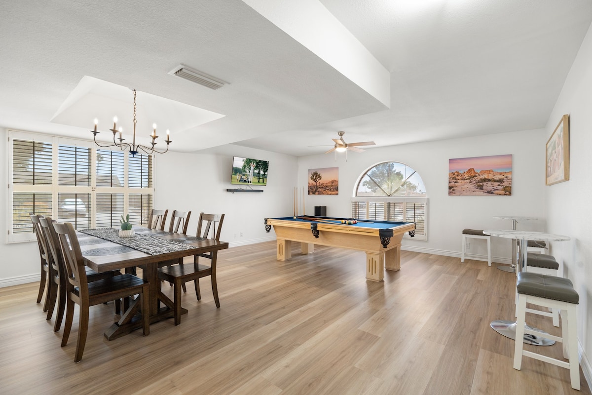 A bright and open indoor living space features a dining table with multiple chairs, a pool table, and a seating area. Large windows allow natural light to illuminate the room, showcasing simple artwork on the walls and a ceiling fan for comfort.
