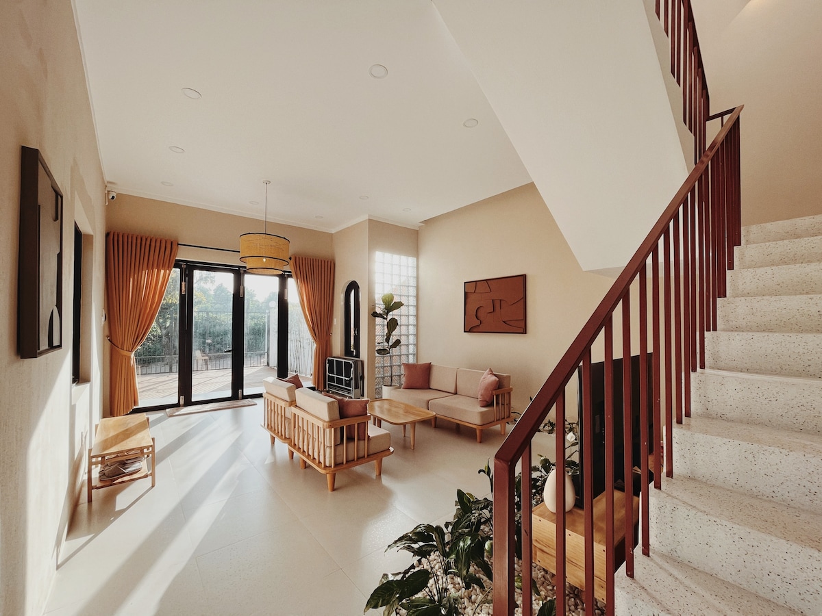 A modern living area is highlighted by large windows allowing natural light to fill the space. Comfortable seating arrangements include a low-profile sofa and wooden chairs. A statement lighting fixture hangs centrally, while indoor plants add a touch of greenery. The staircase leads to an upper level.