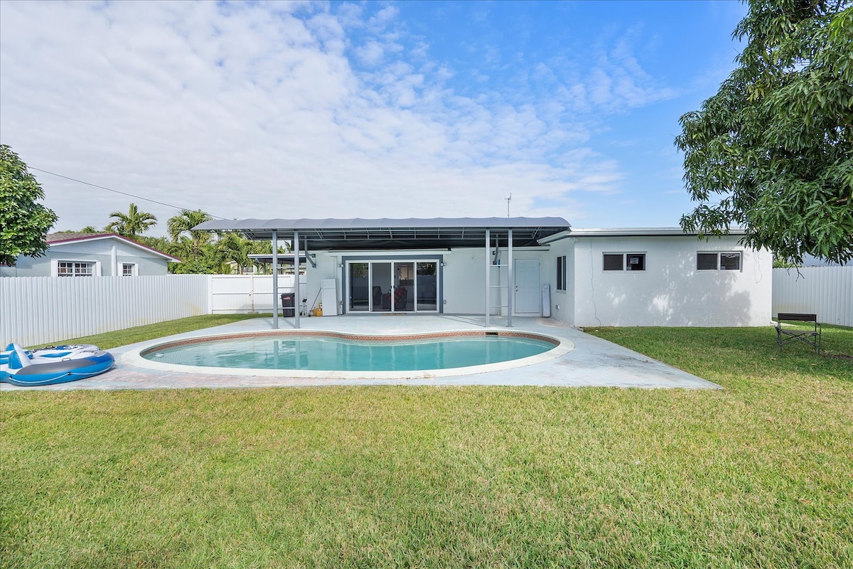 A newly renovated home features a spacious outdoor area with a large pool surrounded by a grassy yard. A covered patio extends from the rear of the house, providing shade. The fenced backyard offers privacy and room for relaxation or play.